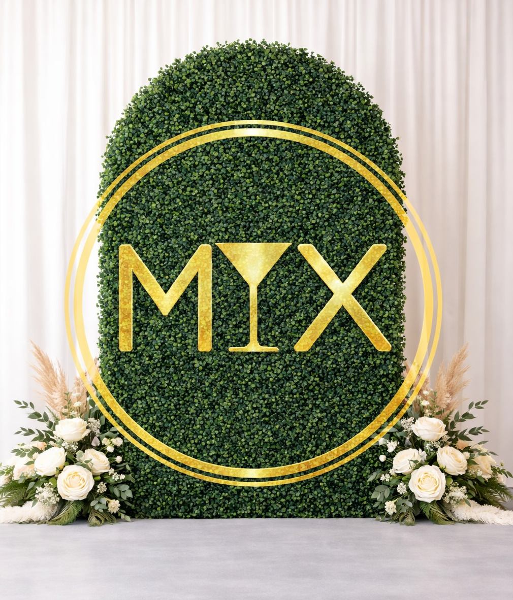 A lush green hedge backdrop with "MIX" in gold letters and a martini glass symbol, framed by golden circles. White roses and pampas grass adorn the base.